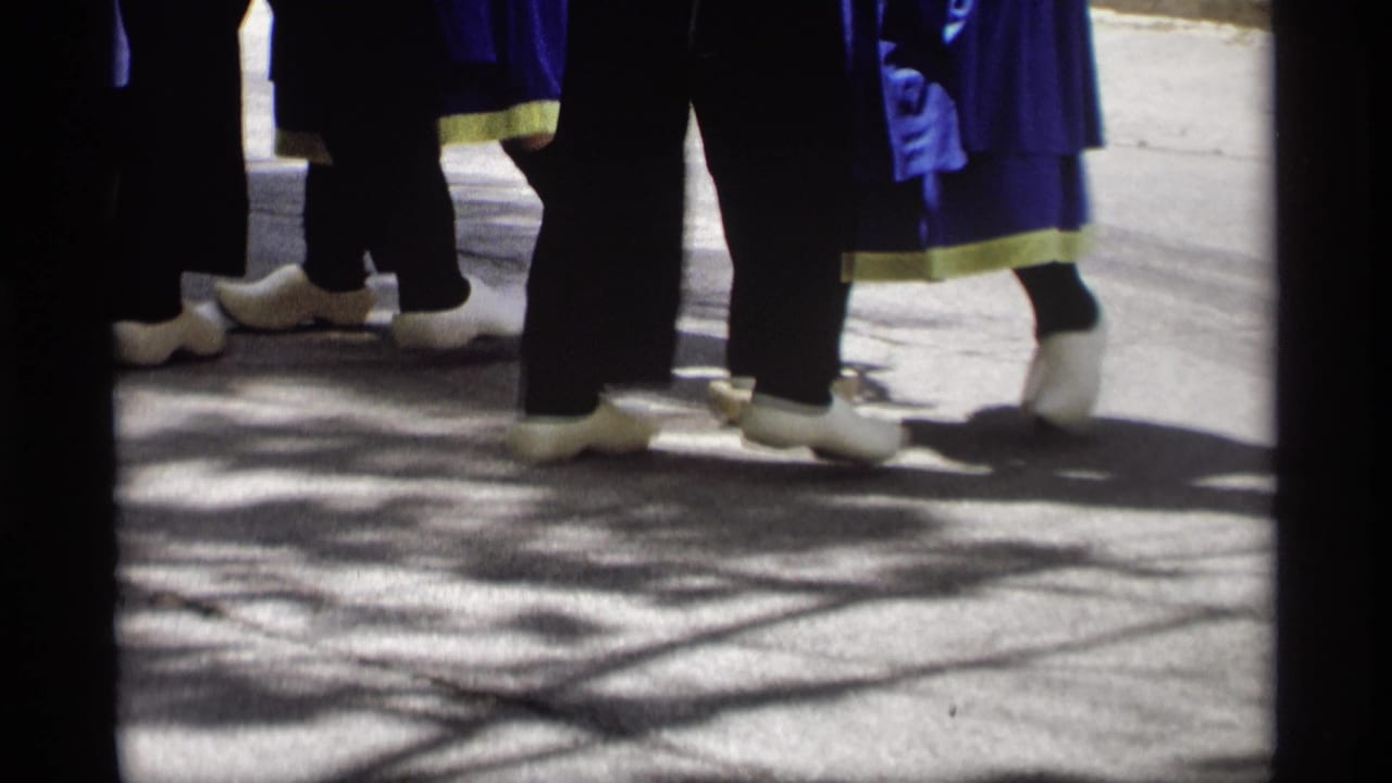 Close view of dancers' legs and white wooden shoes on sunlit pavement.