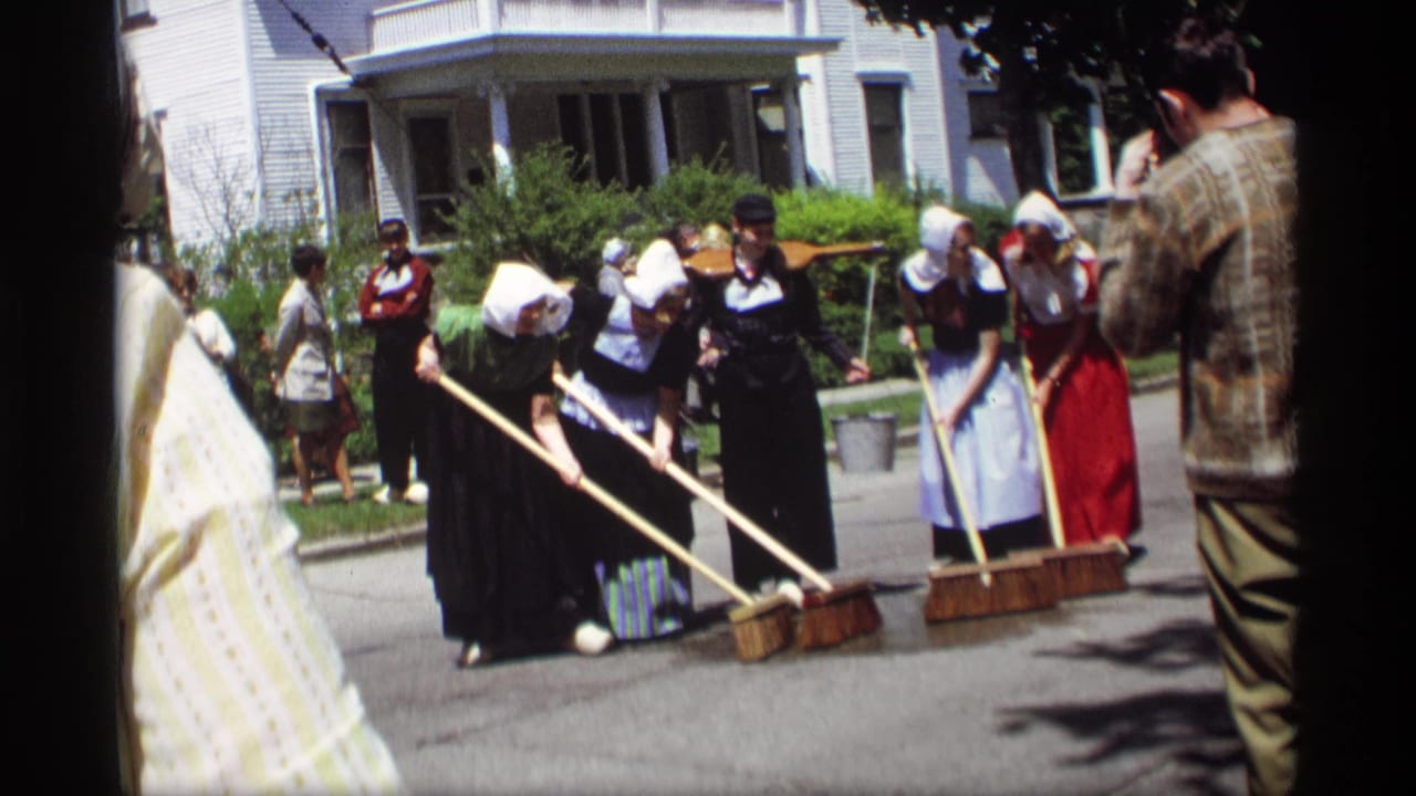 Costumed women sweep a sunlit street with large brooms as spectators watch nearby.