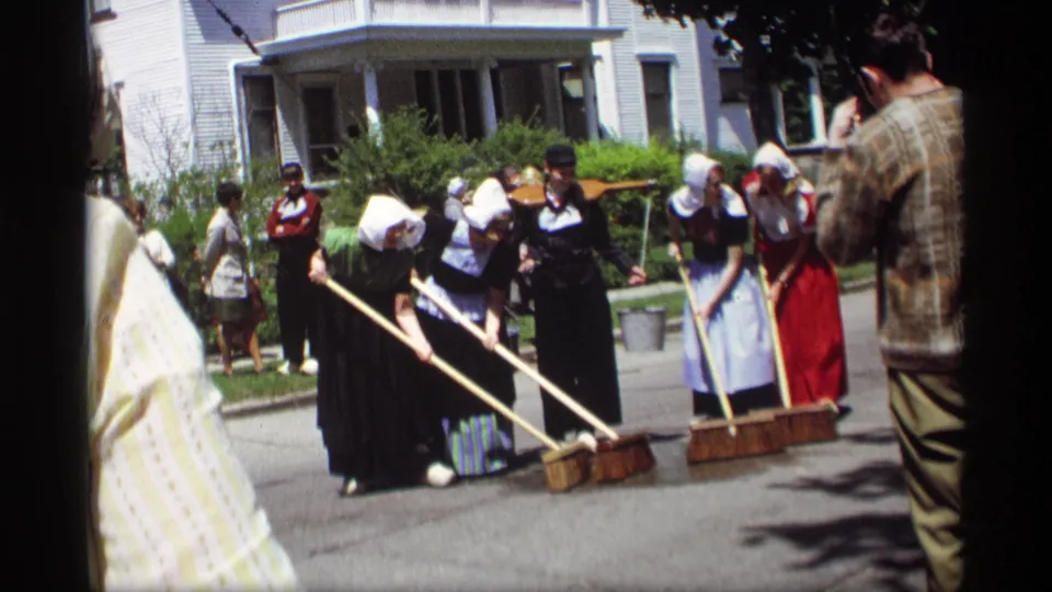 In Holland, Michigan, 1969, women in bonnets, aprons, and long skirts sweep pavement with large brooms while onlookers stand close by.