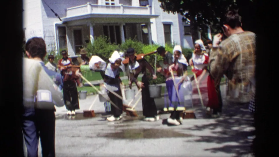 A wider 1969 Holland, Michigan frame shows costumed women using brooms and buckets on wet pavement as spectators watch and film.