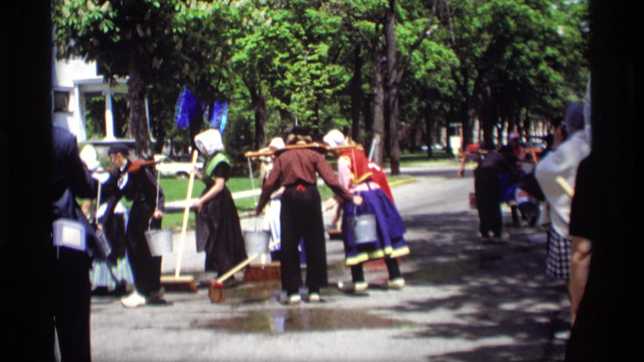 Women in Dutch-style costumes sweep a wet street with buckets nearby.