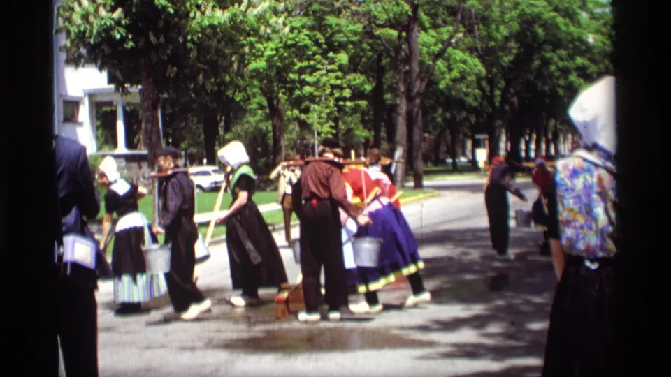 1969 Holland, Michigan frame with Dutch-style folk costumes, brooms, buckets, wet pavement, and spectators along a shaded street.