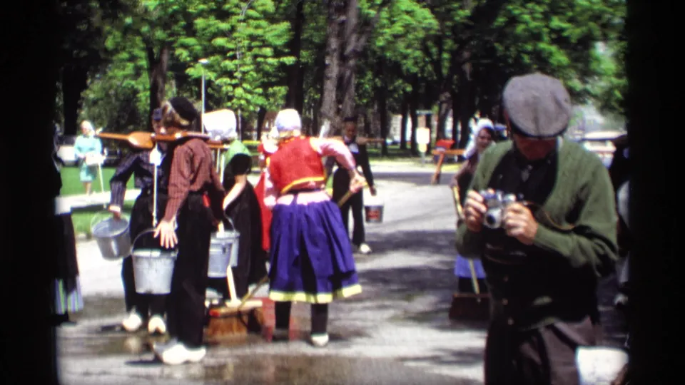 1969 Holland, Michigan home-movie frame showing a camera-carrying man near women in Dutch-style costumes sweeping a wet street.
