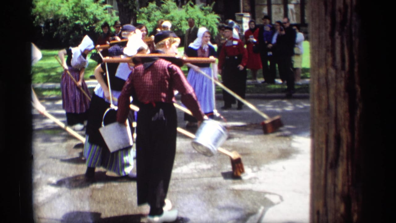 Costumed participants sweep wet concrete with long brooms while spectators watch outdoors.