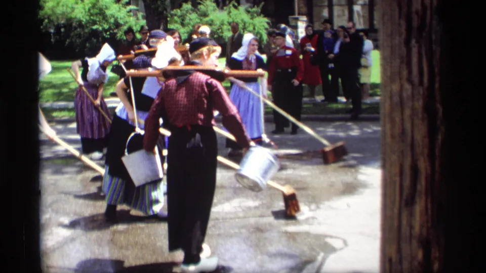 In Holland, Michigan in 1969, people in Dutch-style clothing sweep wet pavement with brooms and buckets as onlookers stand nearby.