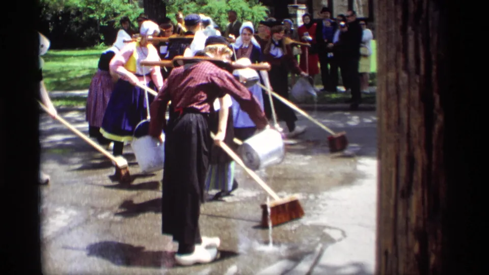 A 1969 Holland, Michigan street-cleaning demonstration shows costumed participants with brooms, buckets, and a shoulder yoke on wet concrete.