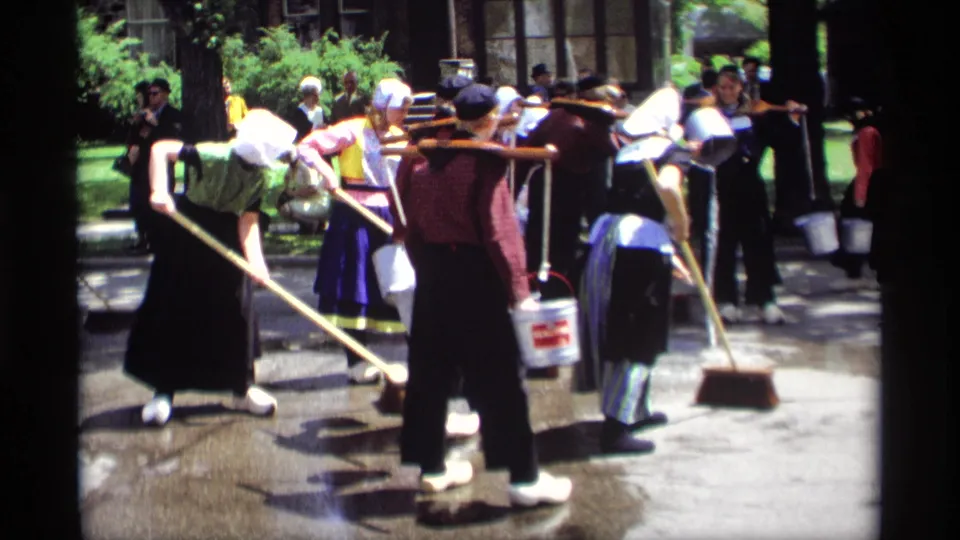 At the end of the clip, Dutch-style costumed participants sweep wet concrete in Holland, Michigan, with onlookers and trees in the background.