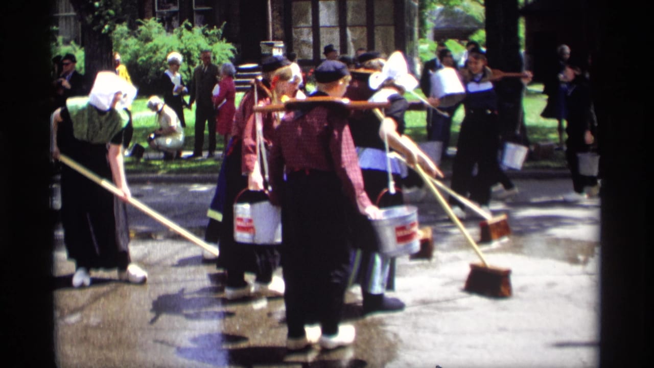 Costumed women sweep a wet street with brooms while carrying metal buckets.