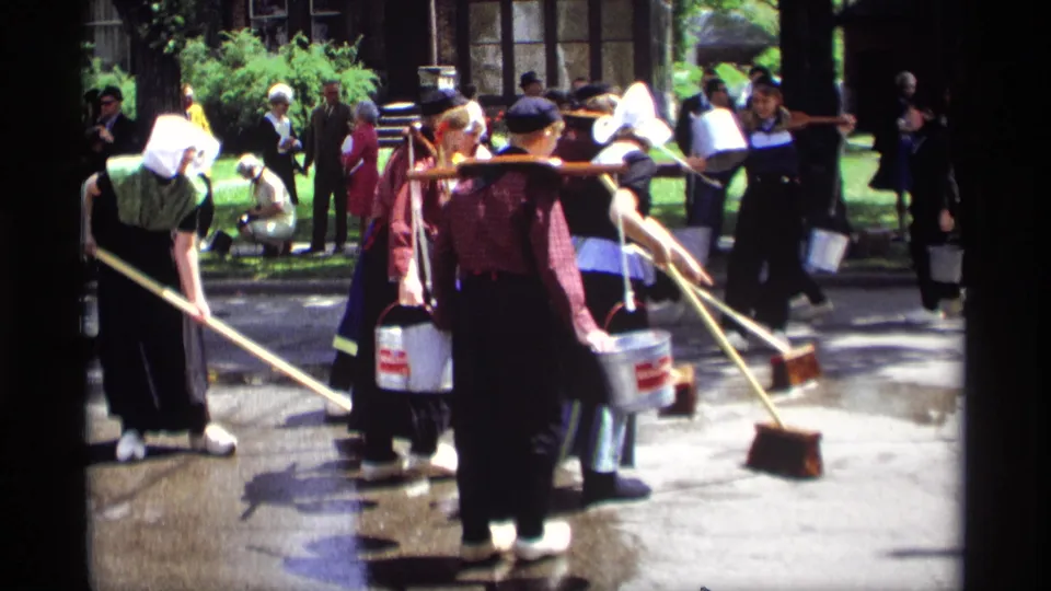 In 1969 Holland, Michigan, women in Dutch-style costumes sweep wet pavement with wooden brooms as onlookers stand near trees and a building.