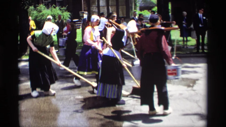 Women in Dutch-style costumes sweep a sunlit street in Holland, Michigan, with buckets nearby and spectators standing along the edge.