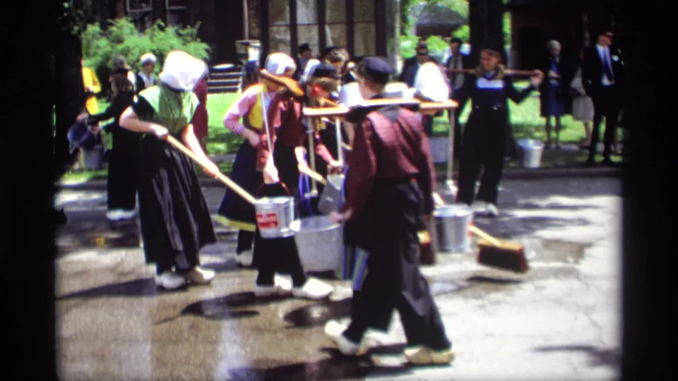 A group of women in bonnets, aprons, and dark skirts sweep and carry metal buckets during a 1969 Holland, Michigan street event.