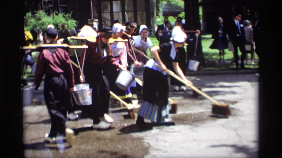 Women in Dutch-style clothing sweep a Holland, Michigan street with long wooden brooms while onlookers and metal buckets remain in the background.