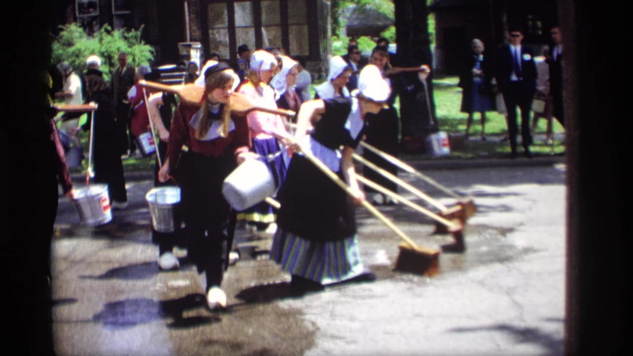 Women in Dutch-style garments sweep wet pavement with long brooms as spectators stand nearby.