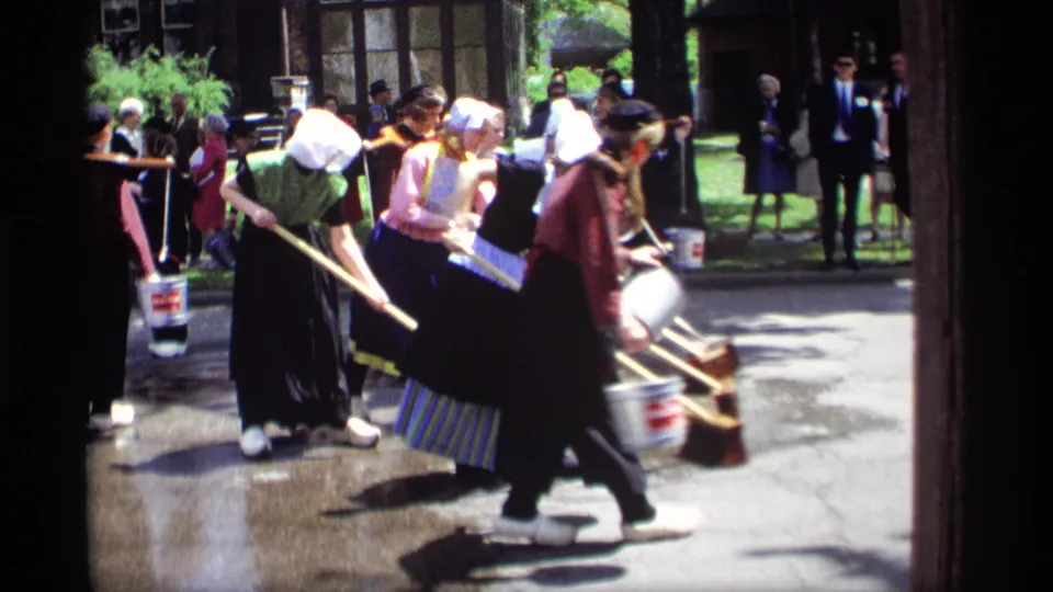 Women in Dutch-style garments move through a Holland, Michigan street-cleaning demonstration with buckets, brooms, and spectators visible.