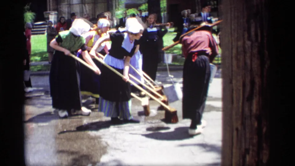 Home-movie frame from Holland, Michigan in 1969 showing women in Dutch-style clothing sweeping while a bucket carrier and spectators remain nearby.