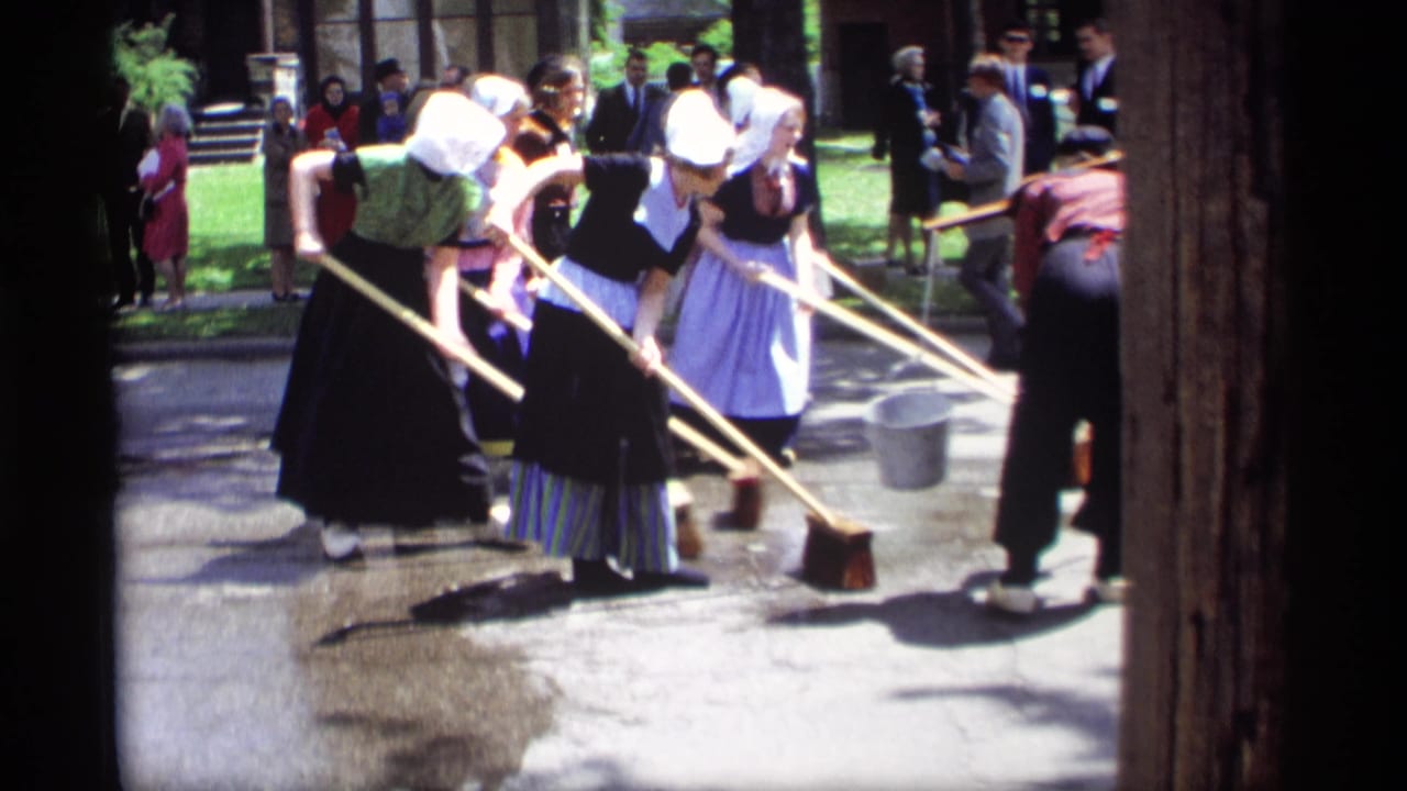 1969 Holland, Michigan home-movie frame showing women in bonnets, dark dresses, and aprons sweeping a wet street while onlookers gather.
