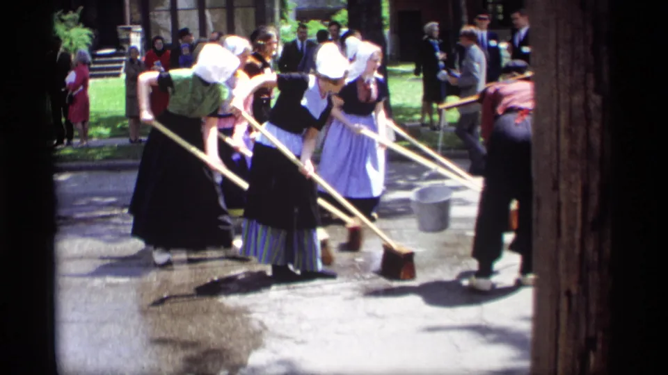 1969 Holland, Michigan home-movie frame showing women in bonnets, dark dresses, and aprons sweeping a wet street while onlookers gather.