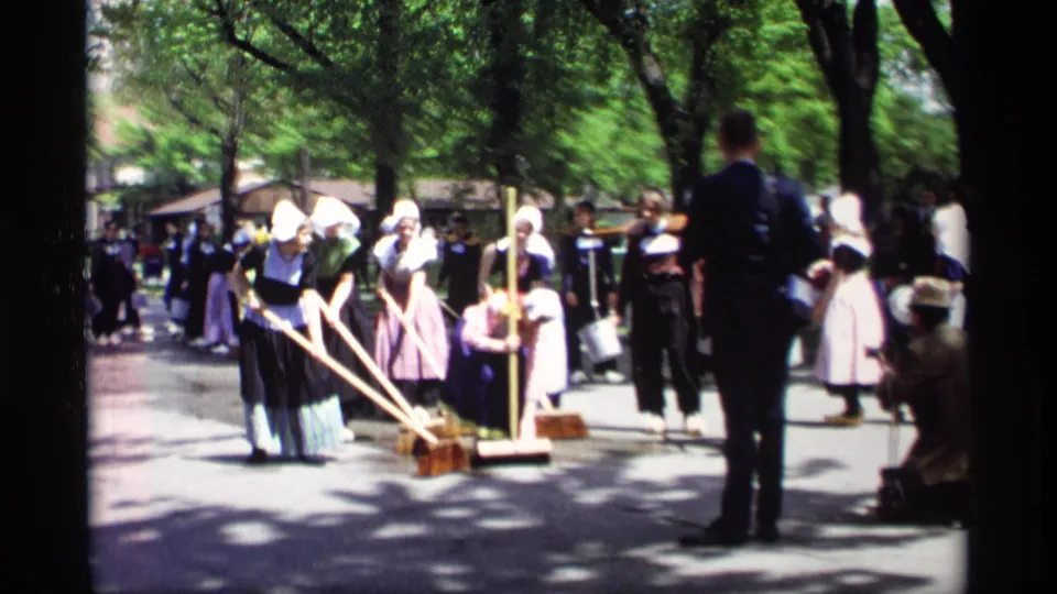 Handheld 8mm view of costumed women sweeping a Holland, Michigan street in 1969, with a man in a suit and spectators nearby.