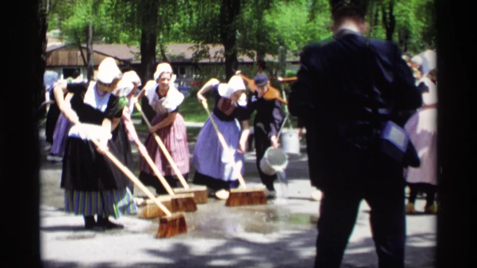 Close street-level frame from 1969 Holland, Michigan showing bonneted women sweeping wet pavement with long brooms during a public gathering.