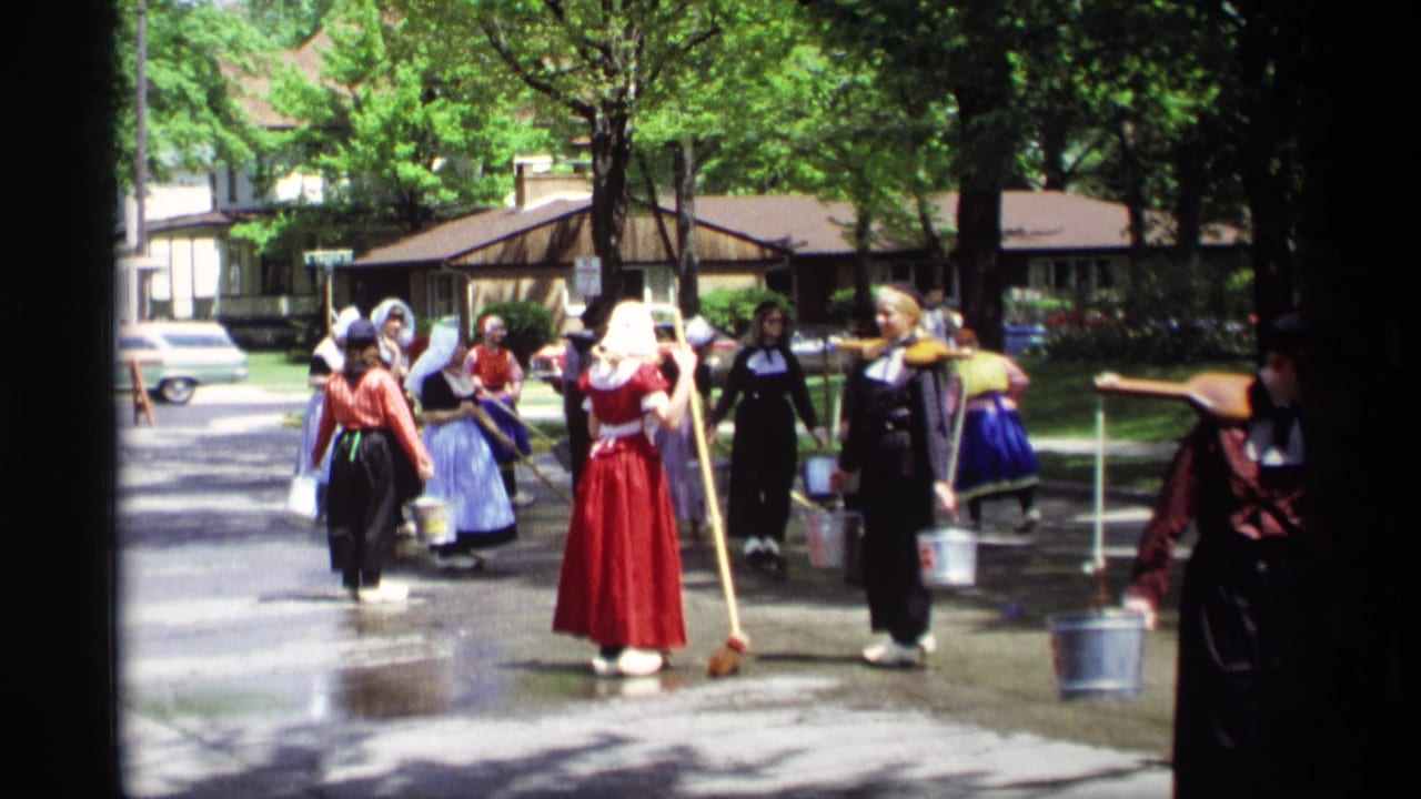 Costumed women with bonnets, skirts, brooms, and buckets walk along a tree-lined residential street.
