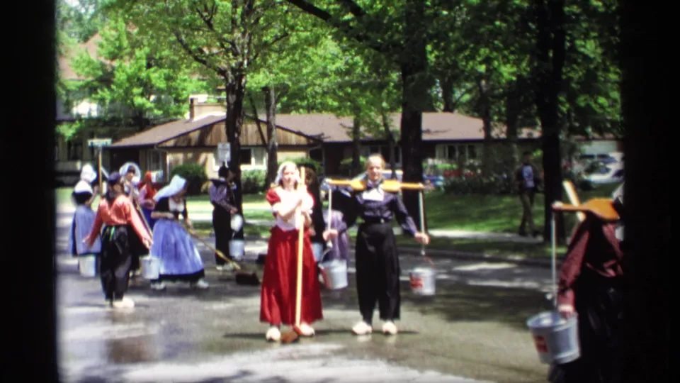 A 1969 Holland, Michigan parade view shows costumed women on a wet street, with one participant balancing a wooden yoke and pails.