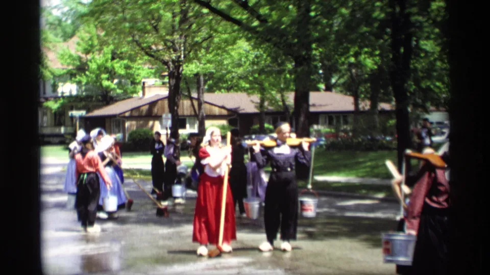 Women in Dutch-style parade costumes move through a Holland, Michigan residential street in 1969, with brooms, buckets, and yokes visible.