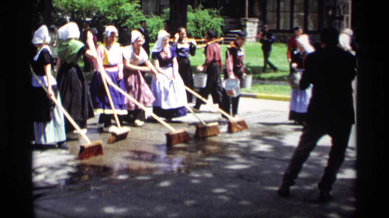 Women in Dutch-style costumes sweep wet pavement with long brooms as onlookers stand nearby.