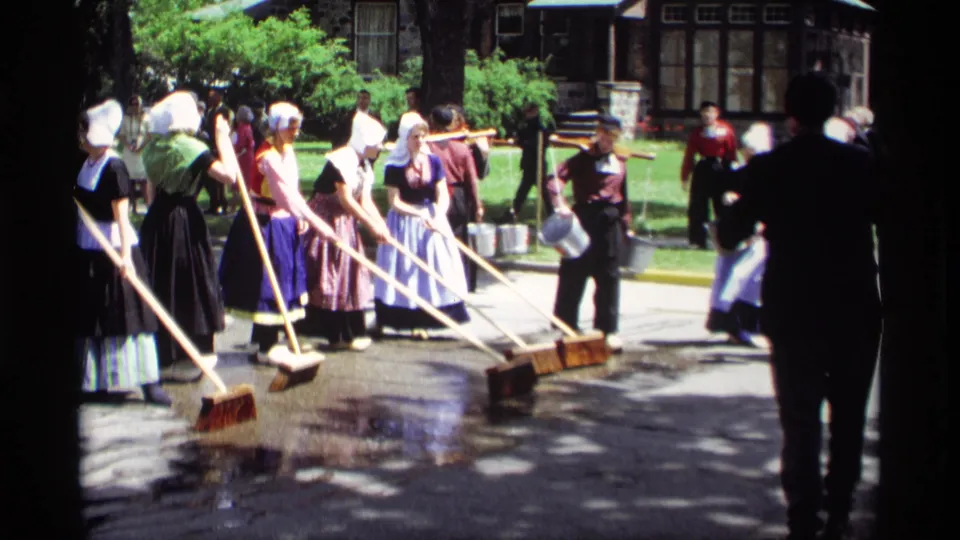 Women in traditional Dutch-style dress continue washing a Holland, Michigan street with brooms and buckets as a passerby crosses the frame.