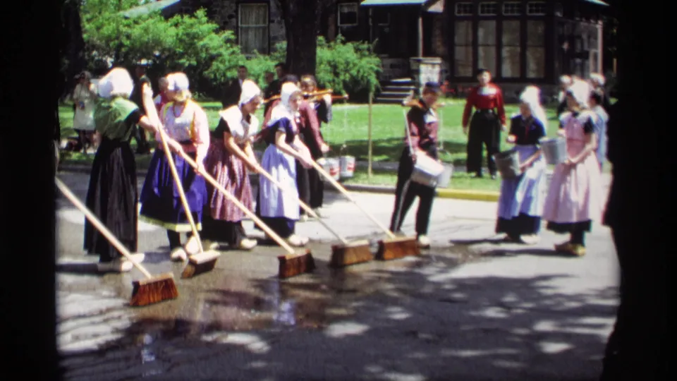 Home-movie view of women in Dutch-style folk costumes pausing and sweeping near buckets on a sunlit street in Holland, Michigan.