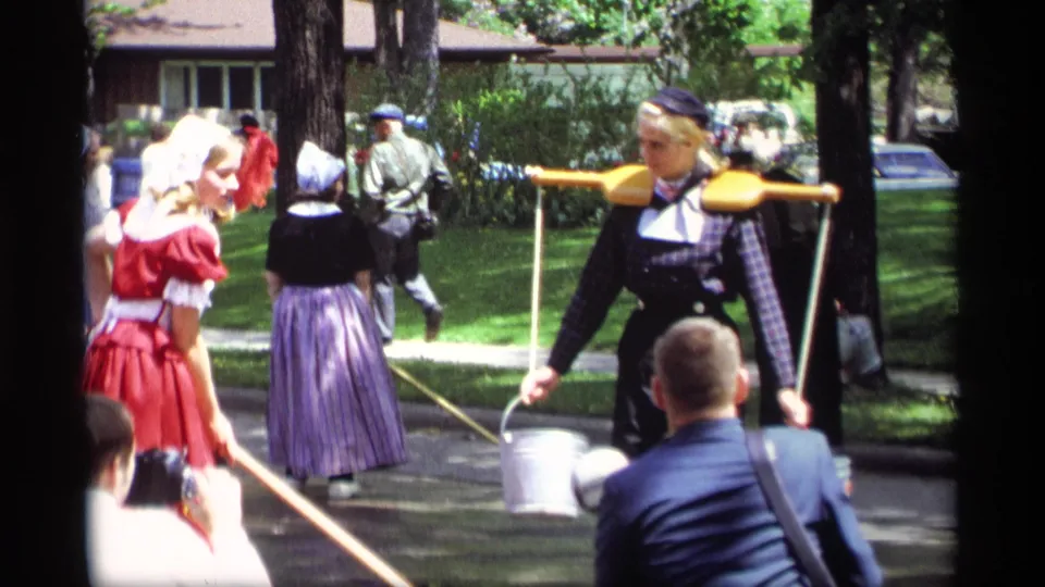 In Holland, Michigan in 1969, women in Dutch-style costumes move past spectators with a wooden yoke, metal buckets, and long poles.