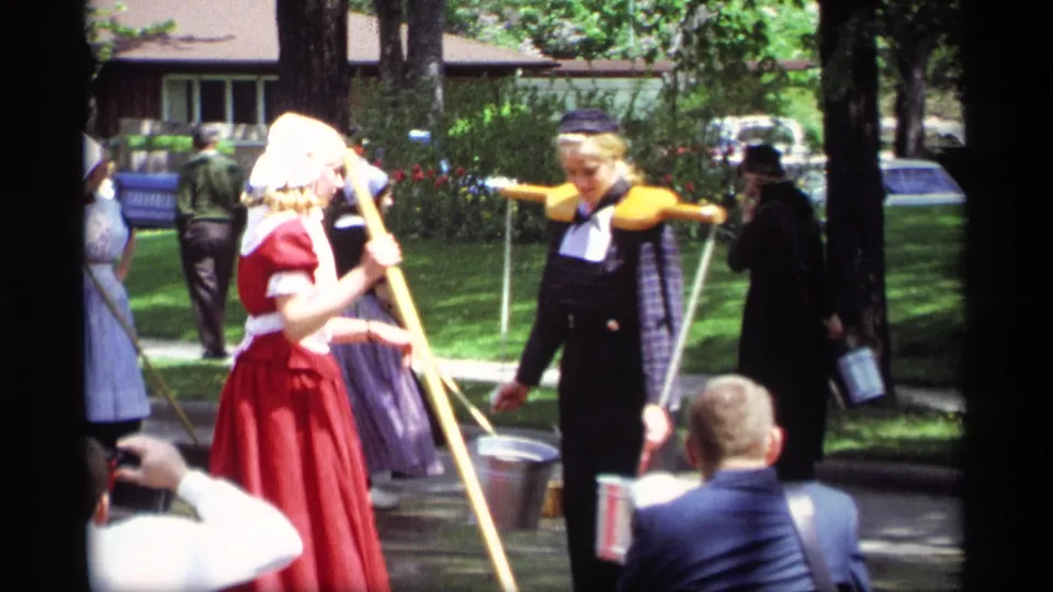 A woman in a red dress and white bonnet walks with a pole as another costumed woman carries a wooden yoke and buckets at the 1969 Holland, Michigan festival.