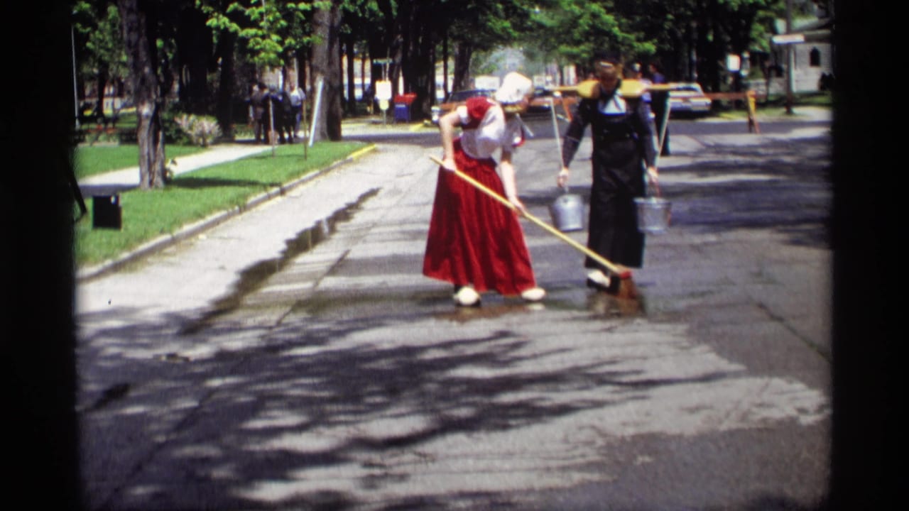 Two women in traditional Dutch-style clothing wash a wet residential street with a broom and bucket.