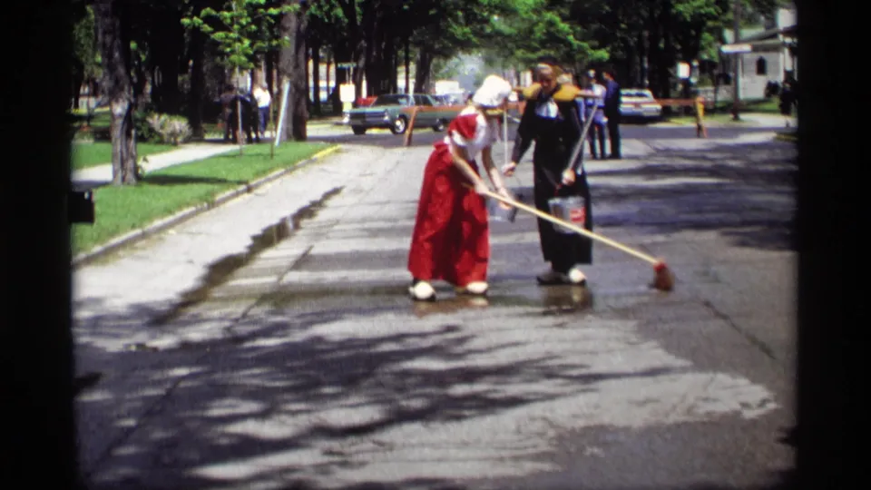 The 1969 Holland, Michigan home-movie frame shows women in Dutch-style dress sweeping water on a residential street.