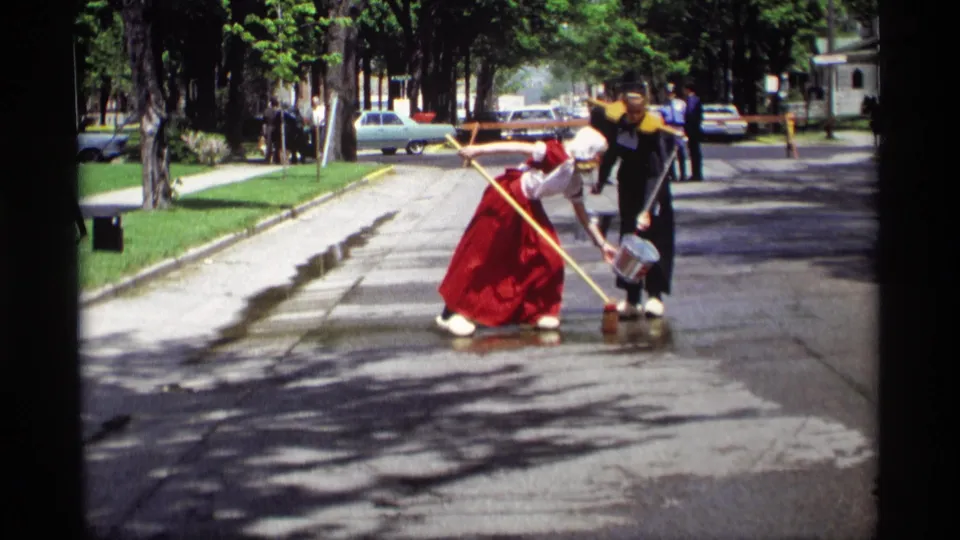 A woman in a red skirt and bonnet bends down during street washing as her companion stands with a bucket in 1969 Holland, Michigan.