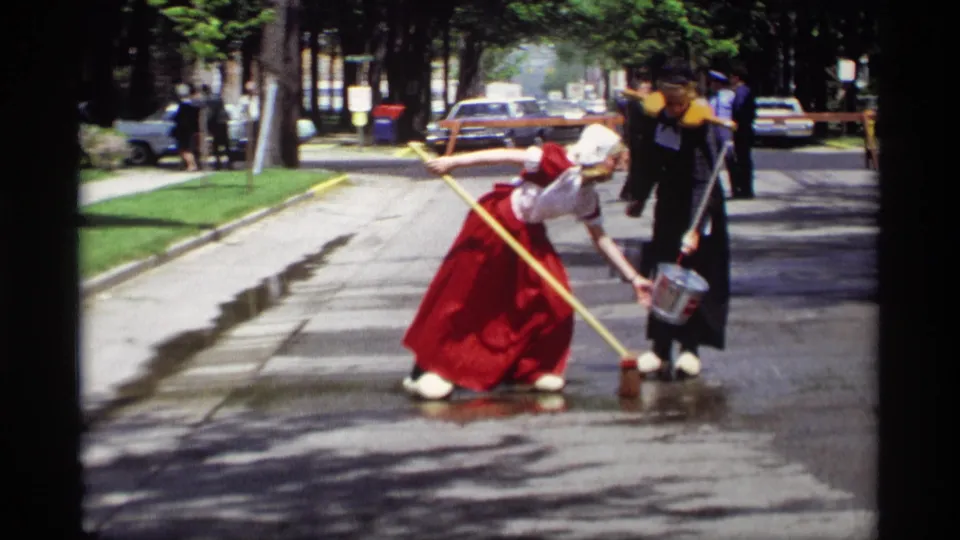 Two women in Dutch-style attire continue washing a Holland, Michigan street, with a bucket, broom, parked cars, and tree shade visible.