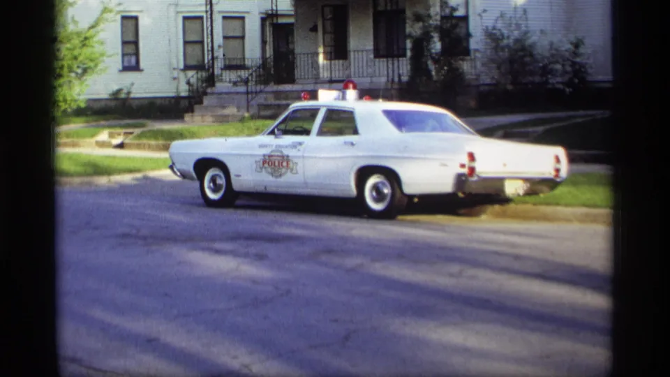 1969 Holland, Michigan 8mm frame showing a white marked police sedan with red roof lights parked on a quiet residential street.