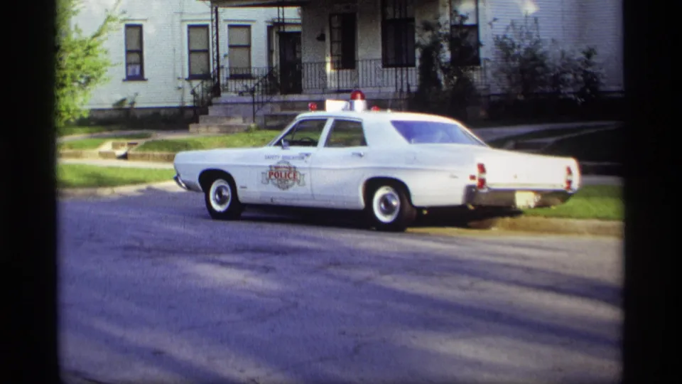 1969 Holland, Michigan 8mm frame showing the side of a white police car parked along a sunlit residential street.