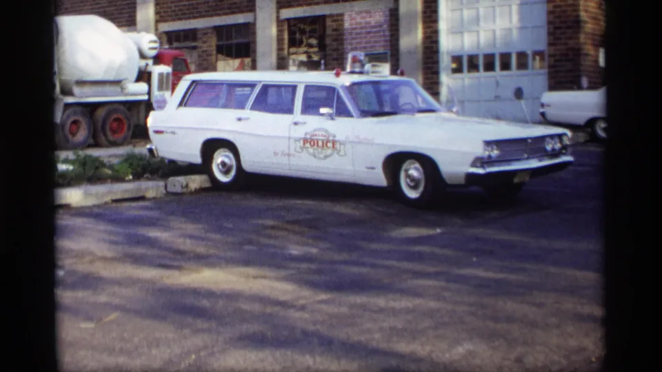 White Ford police station wagon with roof lights sits near a brick building and garage doors in the 1969 Holland, Michigan clip.