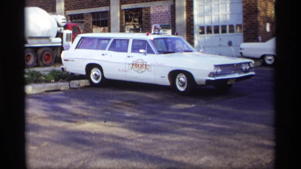 A white Ford police station wagon fills the frame, with door markings, roof lights, brick wall, and garage doors visible behind it.