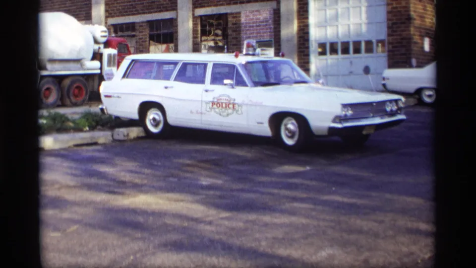 The police station wagon remains parked in the lot, framed with a brick building, garage doors, and part of a nearby truck.