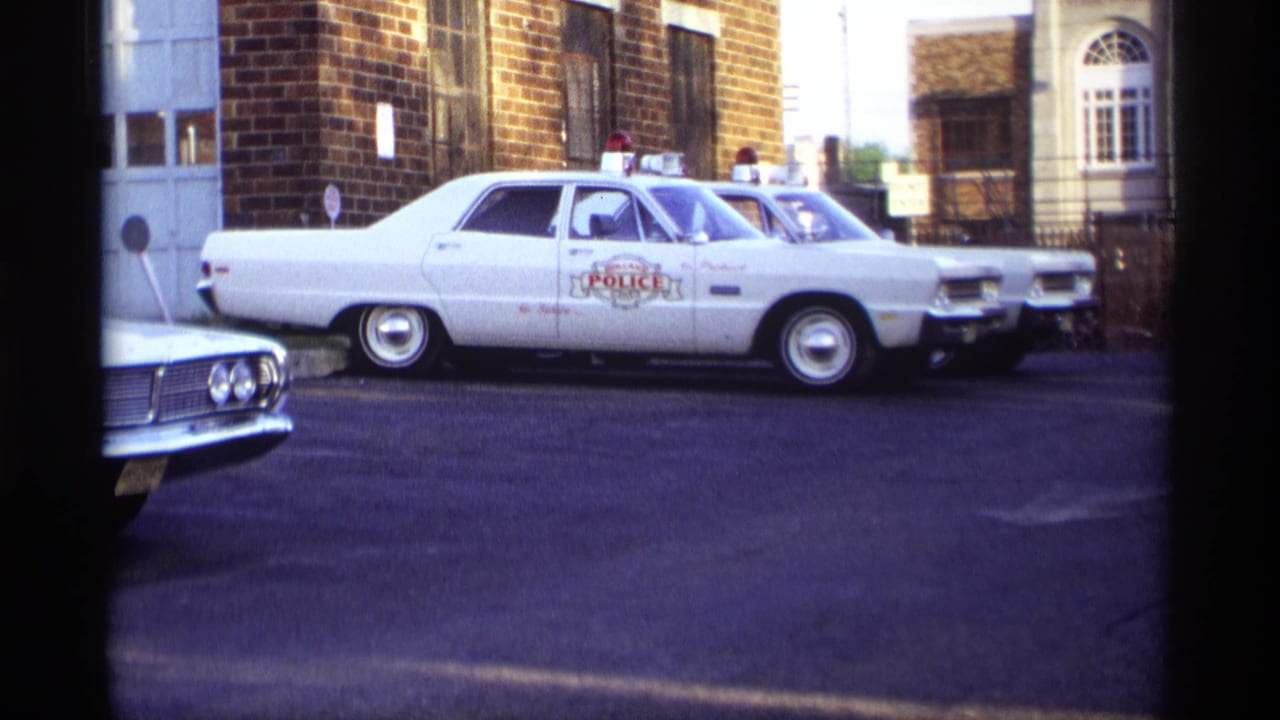 1969 Holland, Michigan street-level frame showing a marked white police car with roof lights parked in a row beside a brick building.