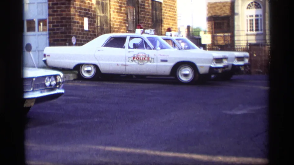 1969 Holland, Michigan side view of a white marked police vehicle parked with other police cars beside brick walls and a gated area.