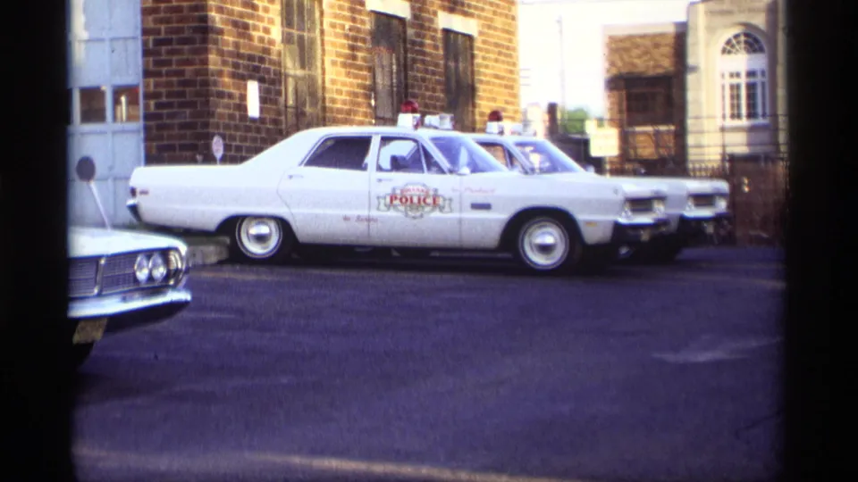 1969 Holland, Michigan frame of a white marked police car and additional police vehicles parked outside a brick building.
