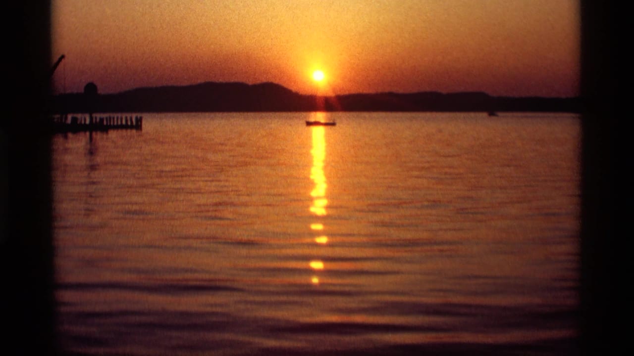 Orange sun setting over a calm lake with a small boat and dark shoreline.