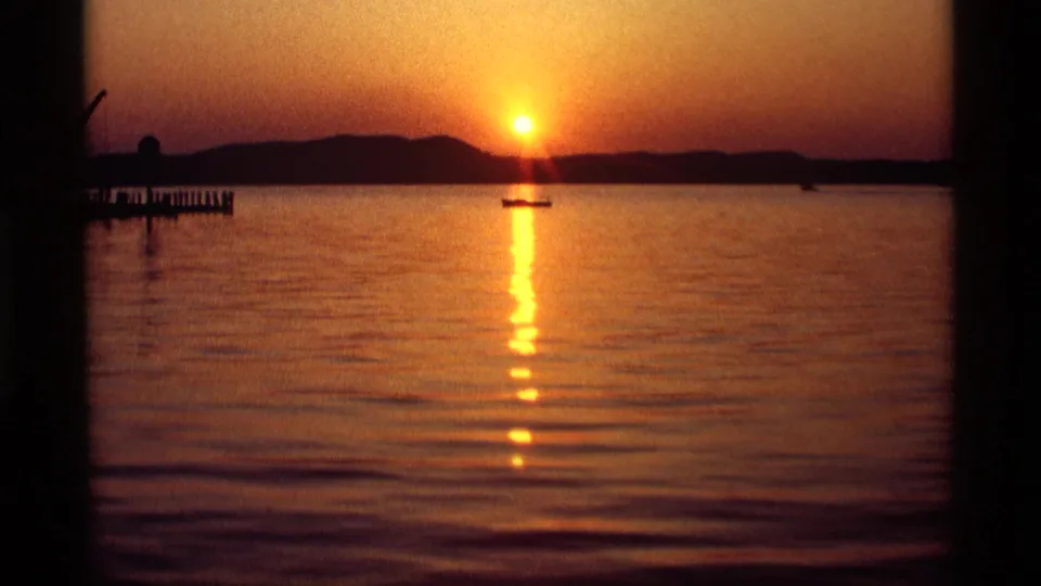1969 Holland, Michigan 8mm frame showing an orange sunset over lake water, with a small boat and distant shoreline in silhouette.