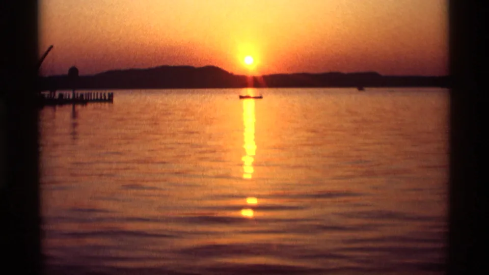 Holland, Michigan lake sunset on 8mm film, with the sun near the horizon, a small boat, and pier posts at left.