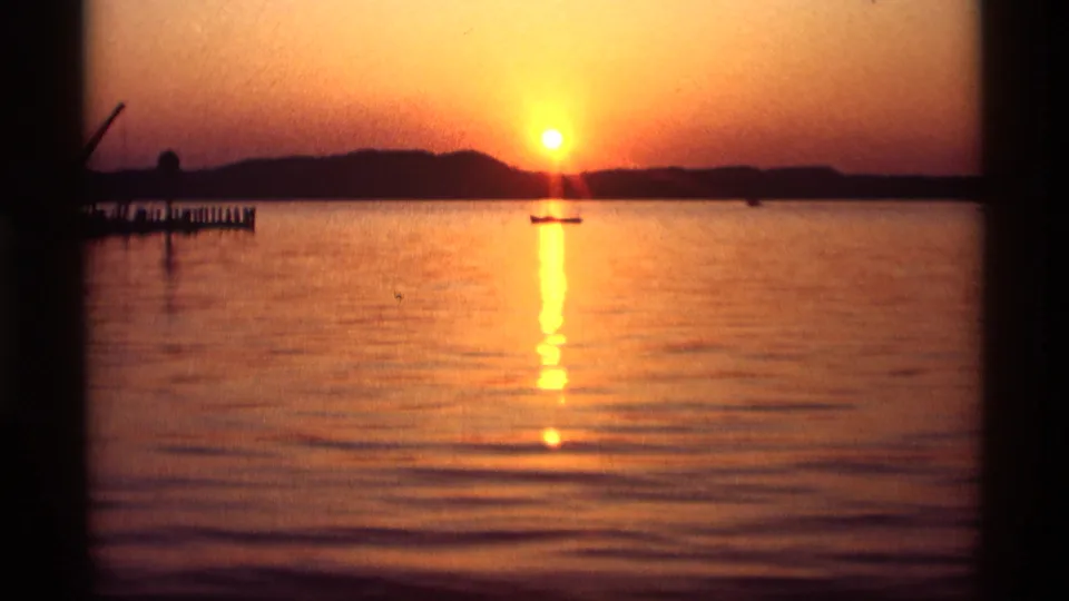 Vintage 8mm view of a Holland, Michigan lake at dusk, with the orange sun near the horizon and a boat centered beneath it.