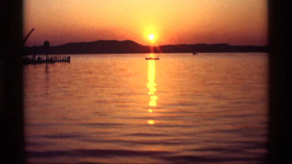 1969 8mm frame from Holland, Michigan showing rippled lake water, a small boat, and the setting sun behind the shoreline.