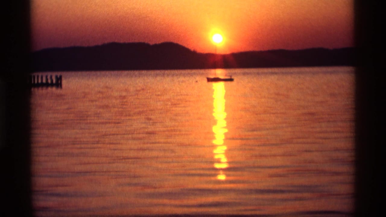 1969 Holland, Michigan lakeside sunset with the sun, dark shoreline, small boat, and shimmering reflection on 8mm film.