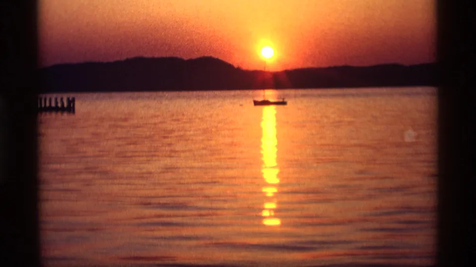 1969 Holland, Michigan 8mm view of a lake sunset, with a silhouetted boat and pier posts against warm orange light.